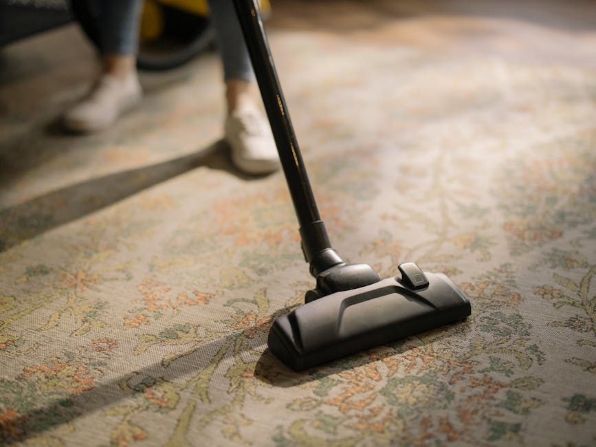 A person holding a yellow portable carpet cleaning machine with a black vent and control panel, positioned over a patterned carpet in a room with exposed brick walls. The machine appears to be used fo